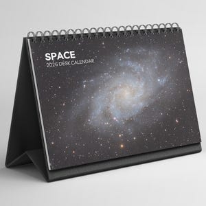 Puede incluir: Un calendario de escritorio negro con encuadernación en espiral, que presenta una imagen de galaxia en la portada. El texto "SPACE 2026 DESK CALENDAR" está impreso en blanco. El calendario está diseñado para colocarse de pie sobre un escritorio.