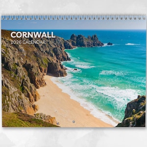 Puede incluir: Calendario de espiral de 2026 con una vista costera de Cornualles. La imagen muestra una playa de arena, agua turquesa y acantilados rocosos bajo un cielo azul. La portada dice "CORNWALL 2026 CALENDAR."