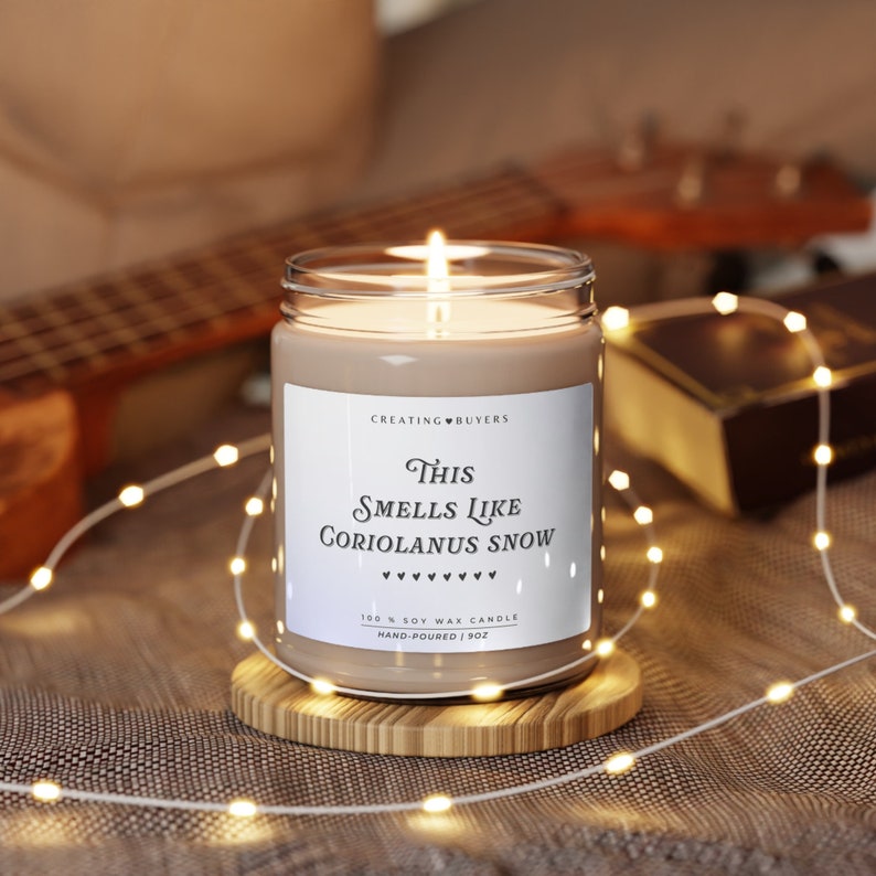 A lit candle in a clear glass jar sits on top of a wooden coaster. The title of the candle is labeled in black against a white background. In the background of the candle is blurred in shades of beige and brown with indistinct items sitting behind the candle.