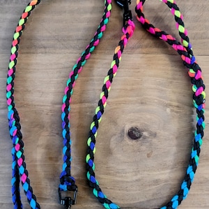 May include: A braided dog leash and matching handle in a vibrant neon color scheme. The leash features a black base with interwoven strands of pink, green, blue, and yellow. A black metal clasp is attached to the leash.