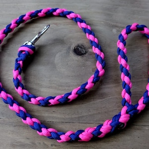 May include: A braided leash in a vibrant pink and blue color scheme. The leash features a silver-colored metal clasp and a loop handle. The rope is tightly woven, creating a durable and stylish accessory for pets.