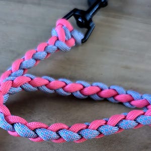 Puede incluir: Una correa de perro trenzada con un esquema de color rosa y azul. La correa est&aacute; unida a un clip de metal negro.