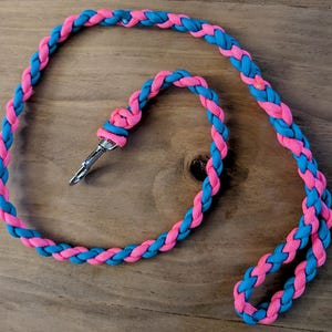 May include: Braided lanyard in pink and turquoise blue. The lanyard is made of a woven cord and features a metal clasp. The cord is braided in a repeating pattern of pink and turquoise blue.