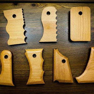 Op de afbeelding: Een verzameling van zeven houten keramiek ribgereedschappen in verschillende vormen. Elk gereedschap is lichtbruin van kleur en heeft een gat om op te hangen. Sommige hebben geschulpte of gebogen randen, andere zijn rechthoekig of schuin.