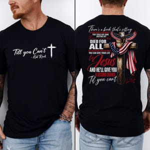 Puede incluir: Una camiseta negra de manga corta con texto blanco en el frente que dice Till you Can't - Kid Rock con un símbolo de cruz. La parte posterior muestra una cruz de madera con una bandera estadounidense, un águila calva y texto sobre la fe.