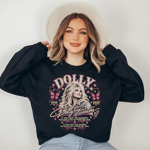 May include: A black long-sleeve sweatshirt featuring a graphic print of Dolly Parton with the text "DOLLY", "1946", "2026", "Dolly Parton", and the quote "If you want the rainbow, you gotta put up with the rain" surrounded by pink butterflies and stars.