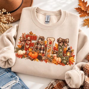 May include: A sand-coloured crewneck sweatshirt featuring the word FALL in large, decorative letters. The letters are adorned with plaid patterns, knit textures, pumpkins, autumn leaves, pinecones, bows, and a small lantern illustration.
