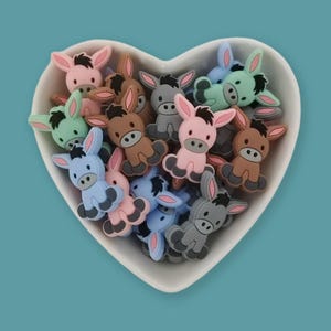 May include: A heart-shaped white bowl filled with colorful donkey-shaped embellishments. The donkeys are in shades of pink, brown, blue, gray, and green. Each donkey has large ears and a small tail.