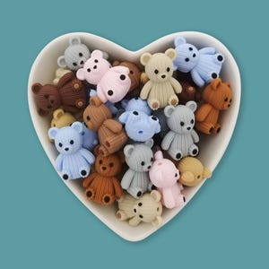 Pode incluir: Uma tigela branca em forma de coração cheia de inúmeras pequenas contas texturizadas em forma de urso em várias cores pastel e neutras, incluindo azul claro, rosa suave, bege, cinza e marrom. Cada conta de urso tem uma superfície canelada.