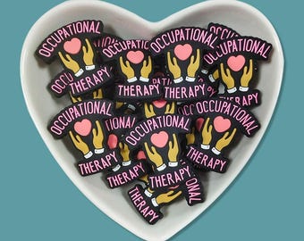 May include: A collection of black and pink Occupational Therapy charms in a white heart-shaped dish. Each charm features a pink heart held by yellow hands, with the words "Occupational Therapy" in pink.