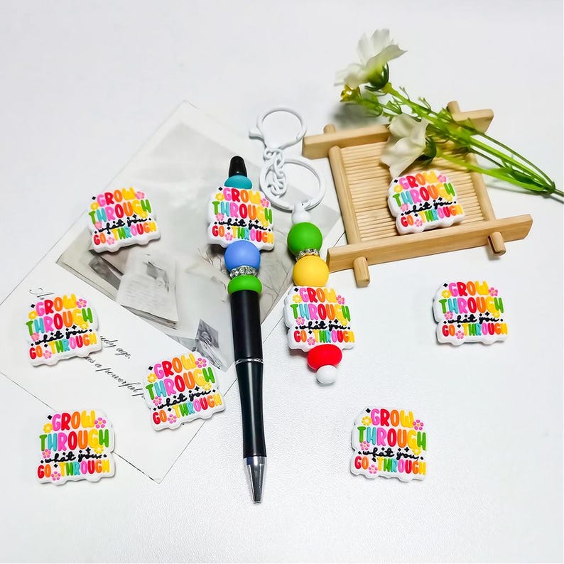 May include: A black pen with a jeweled top and a collection of colorful charms. The charms and pen feature the text "GROW THROUGH WHAT YOU GO THROUGH" in various colors. Additional charms and a keychain are also present.