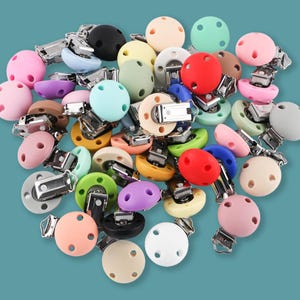 May include: A collection of colorful pacifier clips in various shades, including pink, green, yellow, and blue. Each clip has a round, silicone nipple and a metal clip for attachment. The pacifier clips are scattered on a solid background.