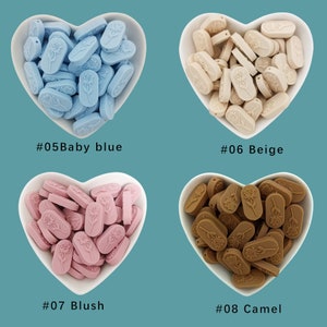 Op de afbeelding: Vier hartvormige kommen gevuld met siliconen kralen in verschillende kleuren: babyblauw, beige, roze blush en camel. Elke kom heeft een nummer en een kleurnaam eronder gedrukt: #05 Babyblauw, #06 Beige, #07 Blush, #08 Camel.