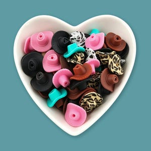 May include: A collection of miniature cowboy hat beads in a heart-shaped bowl. The beads come in various colors including pink, black, brown, turquoise, and leopard print patterns. Each small hat features a central hole for threading or crafting.