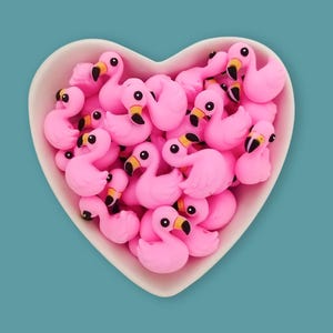 Könnte beinhalten: Eine herzförmige weiße Schale gefüllt mit zahlreichen kleinen, leuchtend rosa Flamingo-Figuren. Jeder Flamingo hat einen gebogenen Hals, einen schwarz-gelben Schnabel und eine glatte, matte Oberfläche, die eine dichte Ansammlung identischer Vogelformen bildet.