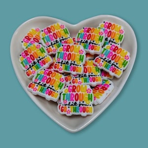 May include: A collection of colorful shoe charms in a heart-shaped dish. Each charm features the words "GROW THROUGH what you GO THROUGH" in a rainbow of colors. The charms are white with a floral design.