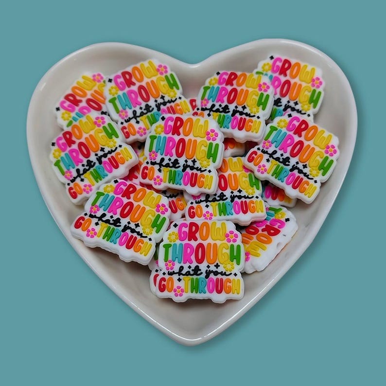 May include: A collection of colorful shoe charms in a heart-shaped dish. Each charm features the words "GROW THROUGH what you GO THROUGH" in a rainbow of colors. The charms are white with a floral design.