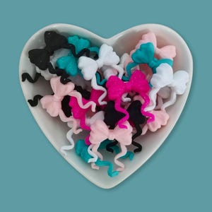 Op de afbeelding: Een verzameling kleine strikvormige bedels in zwart, wit, roze, felroze en talingblauw, elk met twee golvende uiteinden. Deze decoratieve stukken zitten in een witte, hartvormige keramische kom met een gladde, glanzende afwerking.