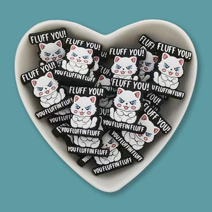Puede incluir: Un bol con forma de corazón lleno de abalorios con forma de gato en blanco y negro. Cada abalorio tiene el texto "FLUFF YOU!" y "YOU FLUFFIN FLUFF" escrito en él.