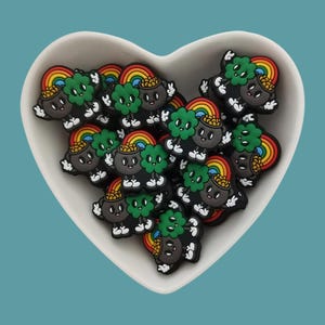 Może przedstawiać: Biała, sercowata miska wypełniona czarnymi, zielonymi i tęczowymi wisiorkami w kształcie garnka ze złotem. Każdy wisiorek ma uśmiechniętą buźkę, koniczynkę czteroliścienną i tęczę.