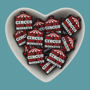 Puede incluir: Un bol lleno de amuletos con forma de carpa de circo roja y blanca con el texto "Not My Circus Not My Monkeys" en cada amuleto.