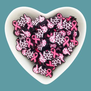 May include: A white heart-shaped bowl filled with pink and black rubber charms. Each charm features a pink ribbon and a hand making the peace sign. The hand is covered in a pink and black leopard print.
