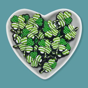 May include: A collection of green and white shamrock-shaped charms with striped and dotted patterns, displayed in a white heart-shaped dish. The charms have black borders and are set against a teal background. Ideal for St. Patrick's Day crafts.