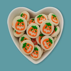 May include: A collection of round, off-white beads featuring a raised orange jack-o'-lantern pumpkin design with a green stem. Each bead has a flat circular surface and is contained within a white heart-shaped bowl, showcasing multiple units.