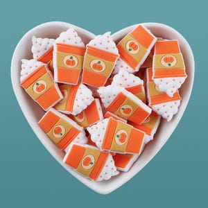 Könnte beinhalten: Eine herzförmige weiße Schüssel gefüllt mit orangefarbenen und beigefarbenen Kürbis-Latte-Charms. Jeder Charm hat eine Kürbisgrafik und eine Schlagsahne-Topping. Die Charms sind verstreut und schaffen ein herbstliches Thema.