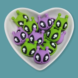 May include: A heart-shaped white dish filled with green and purple alien-shaped embellishments. Each alien has large black eyes and small antennae. The aliens are made of a rubbery material.