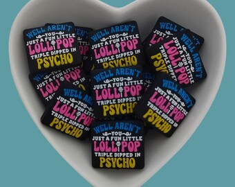 May include: A collection of black, square-shaped charms with the text "Well aren't you just a fun little lollipop triple dipped in psycho" in white, pink, and yellow. The charms are inside a white heart-shaped dish.
