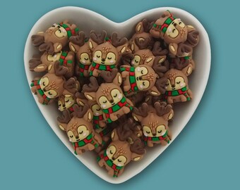 May include: A heart-shaped white bowl filled with multiple small, brown, cartoon-style deer figurines. Each deer has closed eyes, dark brown antlers, and a red and green striped scarf around its neck, creating a festive, repetitive pattern.