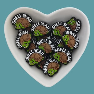 May include: A white heart-shaped dish filled with small, brown and green turtle-shaped charms. Each charm has the words "Shell Yeah" written on it.