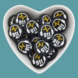 Puede incluir: Un bol blanco en forma de corazón lleno de abalorios redondos de silicona negra y blanca. Cada abalorio presenta un contorno blanco de unos auriculares con una línea EKG amarilla y el número "911" en blanco.