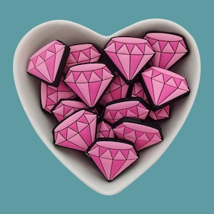Könnte beinhalten: Eine weiße herzförmige Schale gefüllt mit rosa diamantförmigen Silikon-Anhängern.