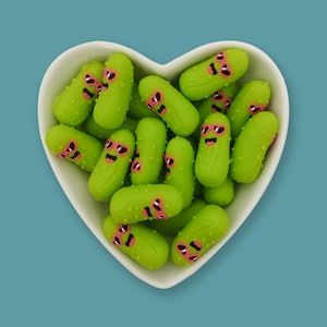 May include: A white heart-shaped bowl filled with numerous small, light green pickle figurines. Each pickle features a textured surface and a printed face wearing bright pink heart-shaped sunglasses, creating a uniform and repetitive decorative display.