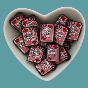 May include: A white heart-shaped bowl filled with pink book-shaped charms. Each charm has the text "SMUT BOOK CLUB" and a red lip design.