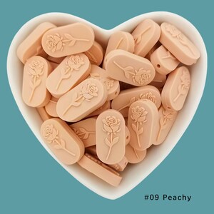 Op de afbeelding: Een kom gevuld met perzikkleurige siliconen kralen, elk met een reli&euml;f van een roos. De kralen zijn ovaalvormig en hebben een gat om te rijgen. #09 Peachy