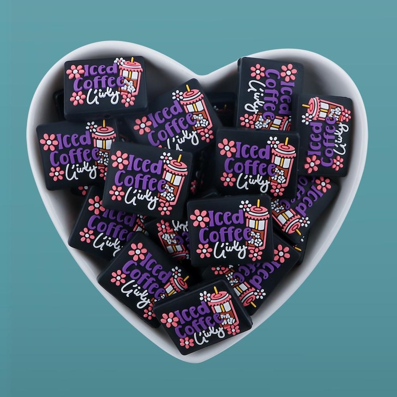 May include: A heart-shaped bowl filled with black and white square-shaped charms. Each charm features a pink and purple cartoon iced coffee cup with the text "Iced Coffee Giuly" in purple.