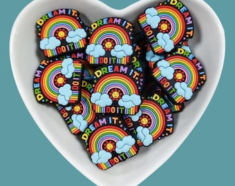 May include: A white heart-shaped bowl filled with colorful rainbow and sun charms. Each charm has the text "Dream It" and "Do It" written on it.