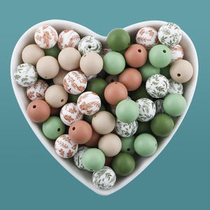 May include: A heart-shaped bowl filled with assorted silicone beads in shades of green, beige, and brown. Some beads have leaf patterns, while others are solid colours. The beads are smooth and round, ideal for crafting.