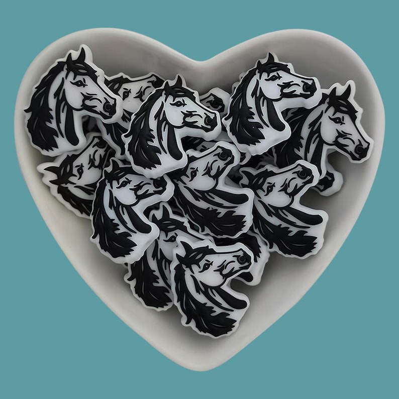 May include: A white heart-shaped dish filled with black and white horse head charms. The charms are made of a smooth, glossy material and have a detailed design.