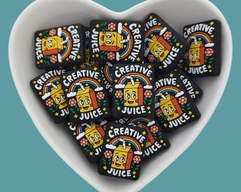 May include: A heart-shaped bowl filled with black square-shaped charms featuring a cartoon illustration of a juice box with a smiling face, a rainbow, and the text "CREATIVE JUICE".