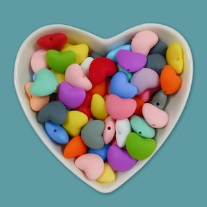 Pode incluir: Uma tigela em forma de coração cheia de contas coloridas em forma de coração em vários tons de vermelho, rosa, amarelo, verde, azul e roxo. As contas são feitas de um material macio e provavelmente são usadas para artesanato ou joias.