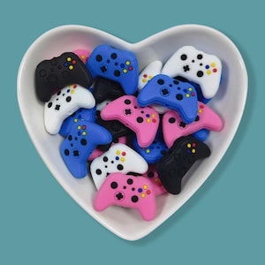 May include: A heart-shaped bowl filled with colorful miniature game controller charms. The controllers are in various colors, including black, white, blue, and pink, with colorful button details. The bowl is white, and the background is a solid teal color.