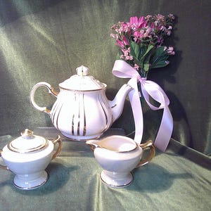 May include: A white teapot with gold trim, a white sugar bowl with gold trim, and a white creamer with gold trim. The teapot has a lid and a spout. The sugar bowl has a lid. The creamer has a handle. All three pieces are on a green surface. A bouquet of pink flowers with a pink ribbon is in the background.