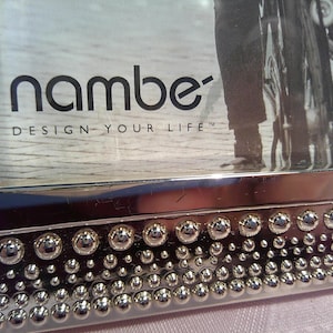 May include: A silver picture frame with a beaded border and a black and white photo of a person riding a bicycle. The photo has the text "nambe" and "design your life" printed on it.
