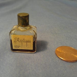 May include: A small, clear glass perfume bottle with a dark brown liquid inside. The bottle has a gold-colored cap and a rectangular label that reads "Réplique, Raphael Paris." A penny is next to the bottle.