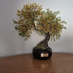 Pode incluir: Uma escultura de árvore bonsai de arame com folhagem de contas amarelas e verdes num tronco castanho texturado. A árvore está num vaso de cerâmica preta brilhante com uma pequena placa dourada na frente com a inscrição "F.LBONSAL".