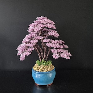 Op de afbeelding: Een handgemaakte draad bonsai boom met een lavendelkleurig bladerdak en een donkerbruine stam. De boom staat in een blauwe keramische pot gevuld met kleine kiezels en groen mos. De bonsai is een decoratief item.